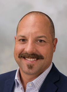 Portrait of a man with glasses and a gray beard wearing a green collared shirt against a neutral background