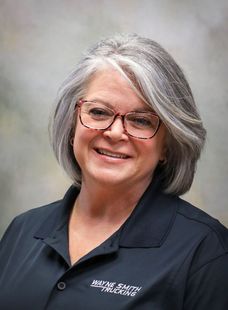 Smiling woman with gray hair and glasses wearing a dark polo against a soft gray background