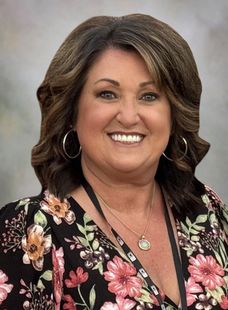 Smiling woman with shoulder-length brown hair wearing a black floral blouse and hoop earrings on a gray background