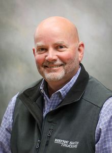 Smiling bald man in a dark vest and plaid shirt against a gray studio backdrop