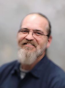 Smiling man with glasses and gray beard against a blurred gray background