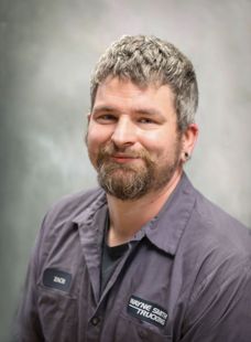Smiling person with short hair and beard wearing a dark work shirt against a gray studio backdrop