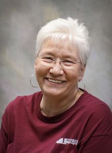 Smiling person with short white hair and glasses wearing a maroon shirt against a gray backdrop