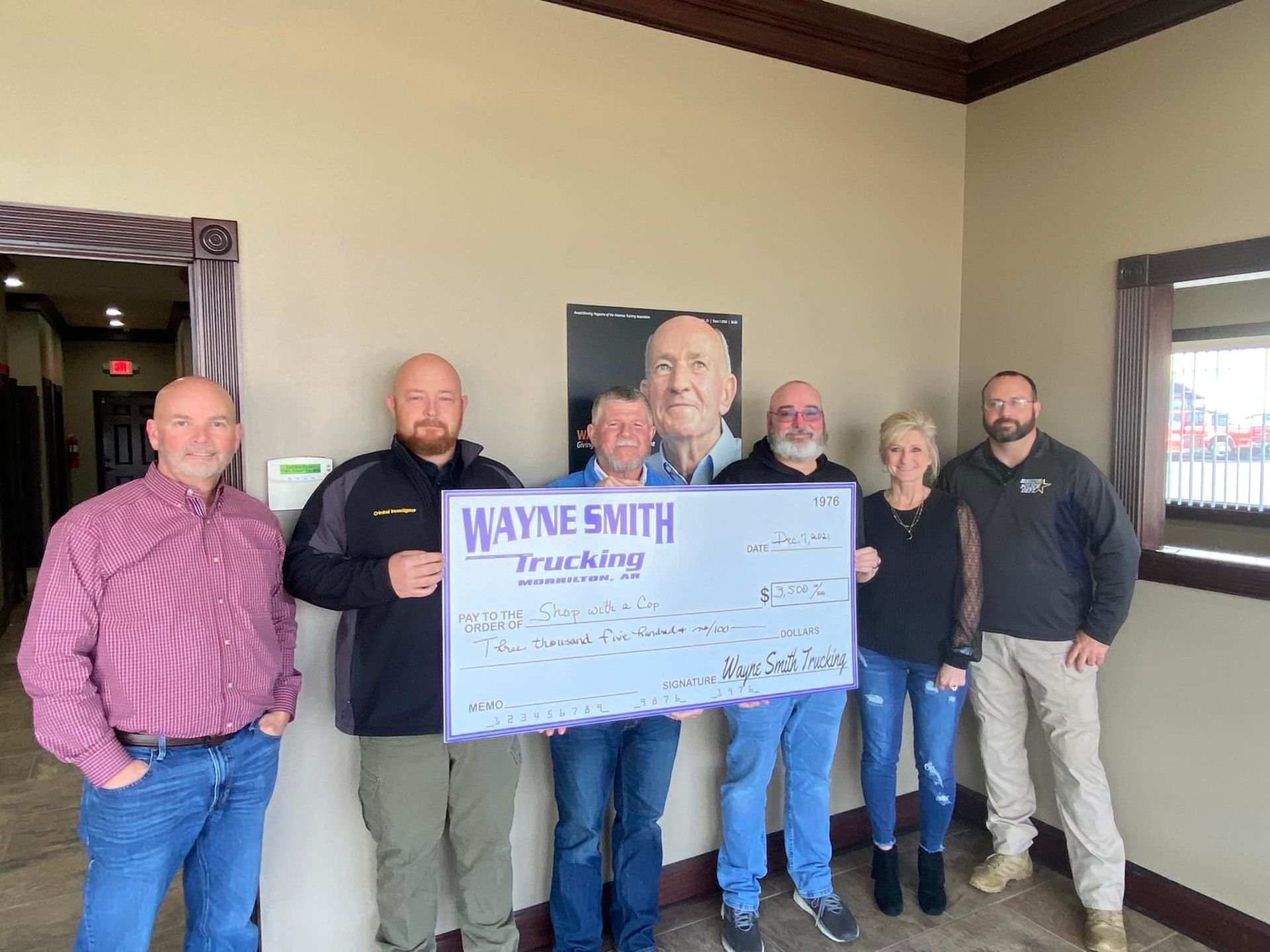 Group of people holding a large check in a room, with a framed portrait on the wall behind them