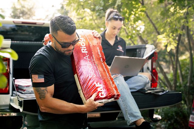 Man holding roofing material, another person using a laptop on a truck bed. Outdoors.