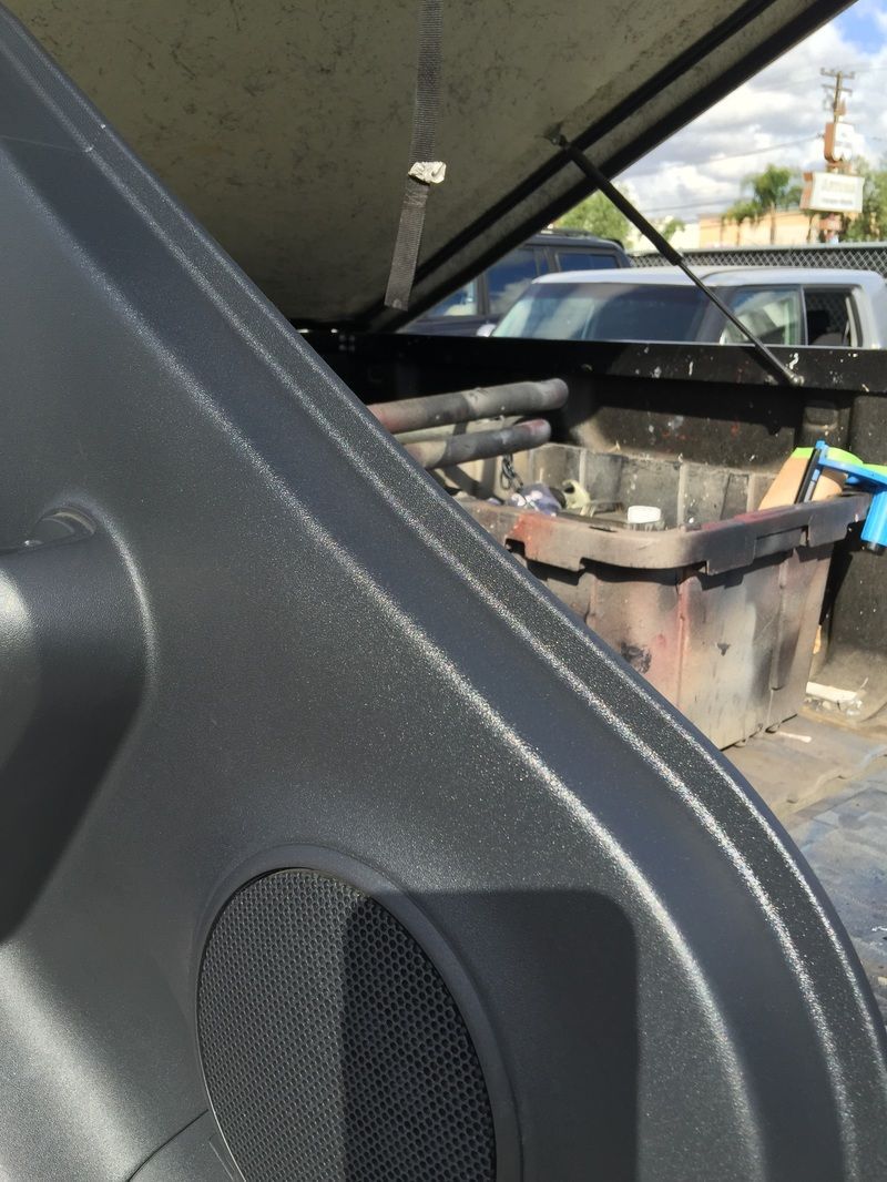 Car’s open hood shows engine components and hoses in a driveway, with a vehicle parked nearby.