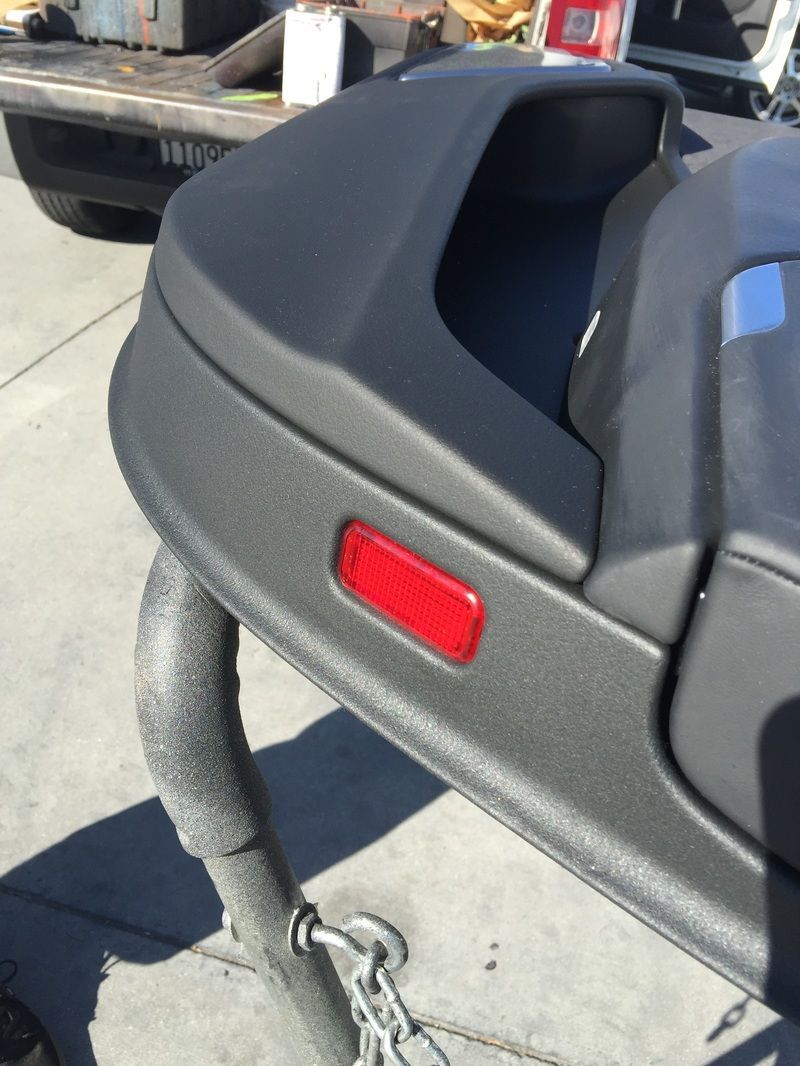 Close-up of a black car bumper with a red reflector and a tow strap attached on pavement