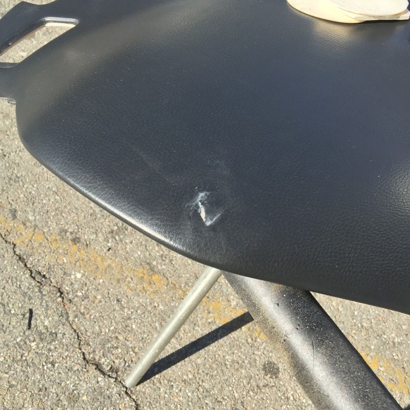 Black chair seat with a small tear, showing metal legs on cracked pavement outdoors