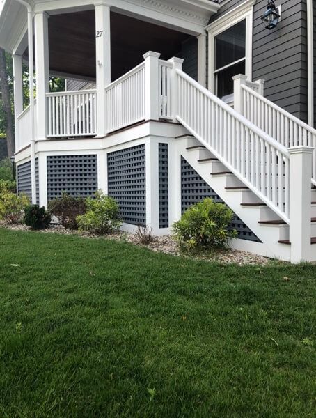 White porch with stairs, lattice skirting, and small bushes on green lawn.
