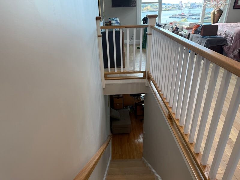 Staircase leading downwards. White walls and railing, wooden handrail, view to lower floor with furniture.