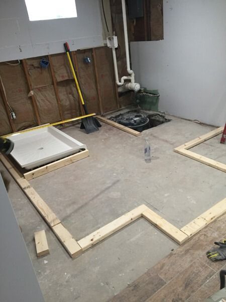 Basement remodel in progress, wooden framing laid out on concrete floor. Shower base and plumbing visible.