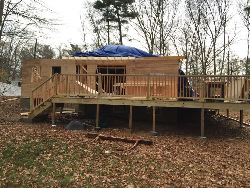 Wooden building under construction with a deck, surrounded by trees and fallen leaves. Blue tarp covers the roof.