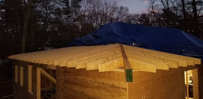 A wooden structure with an unfinished roof. A blue tarp covers a portion of the roof. Dark trees in the background.