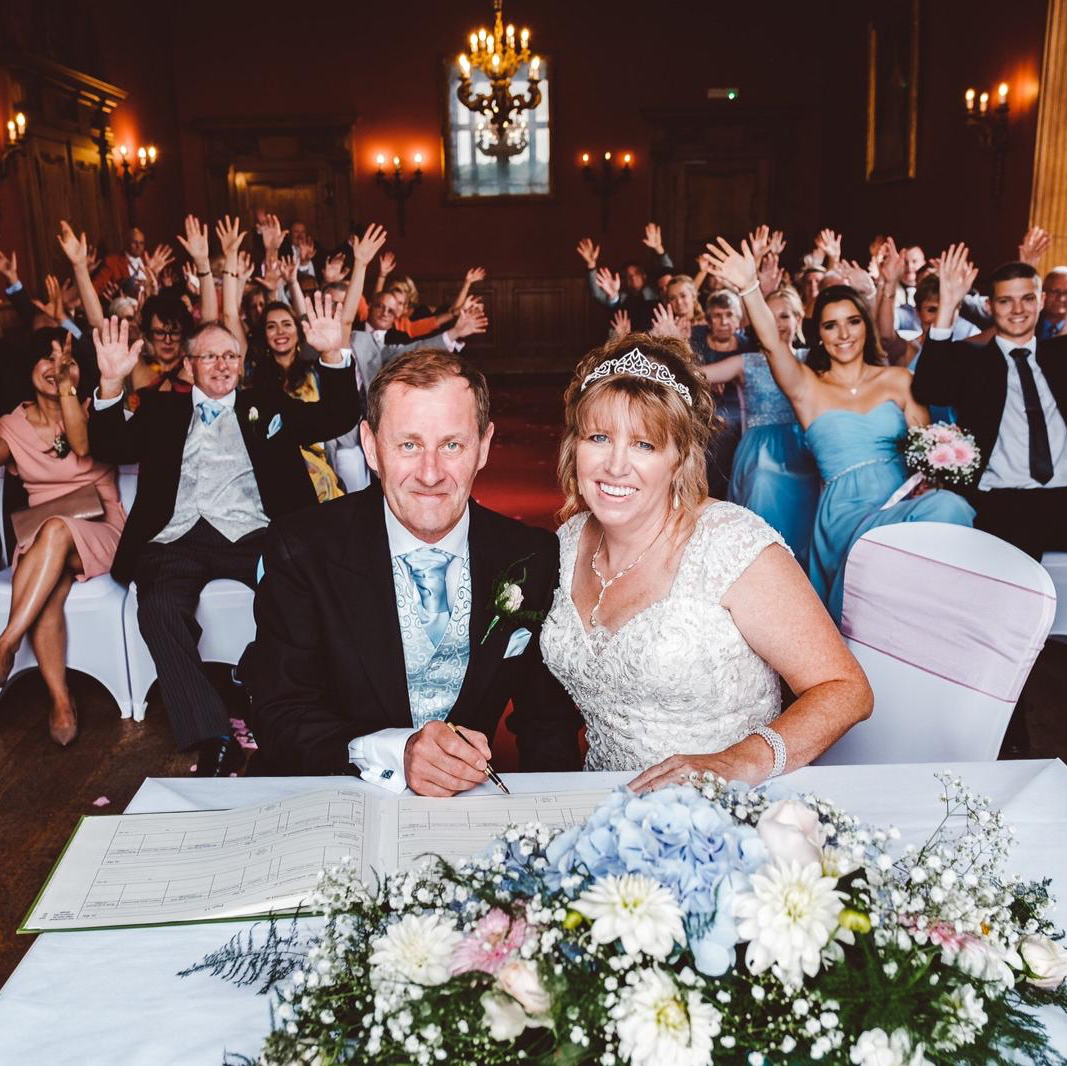 A bride and groom sit at a table with their hands in the air