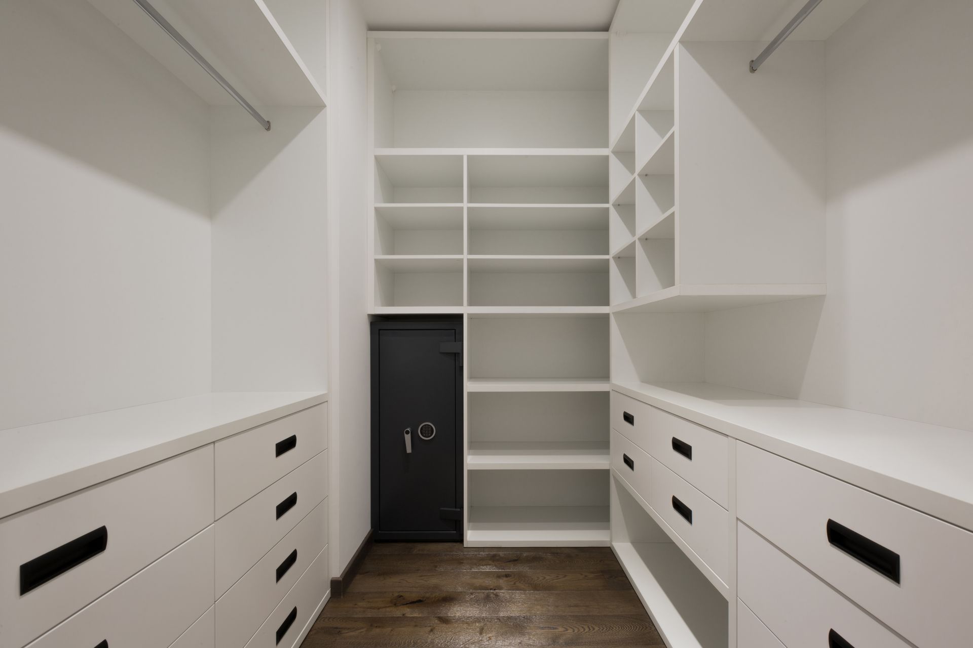 White walk-in closet with built-in shelves, drawers, and dark wood floor, centered on a black safe door.