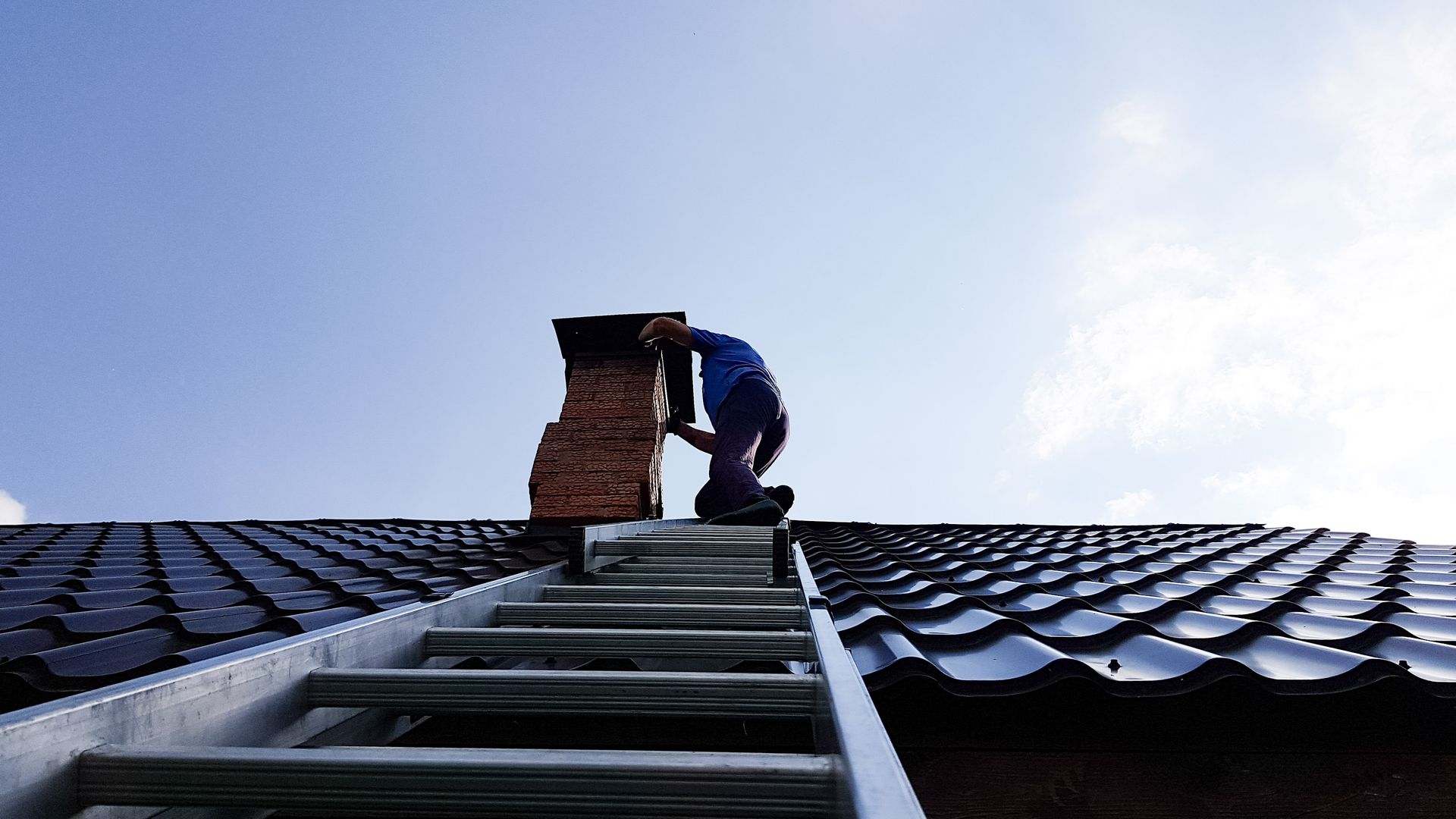 Person on ladder working on a brick chimney on a black tiled roof under a blue sky. Person on a ladder, cleaning a chimney on a rooftop under a blue sky.