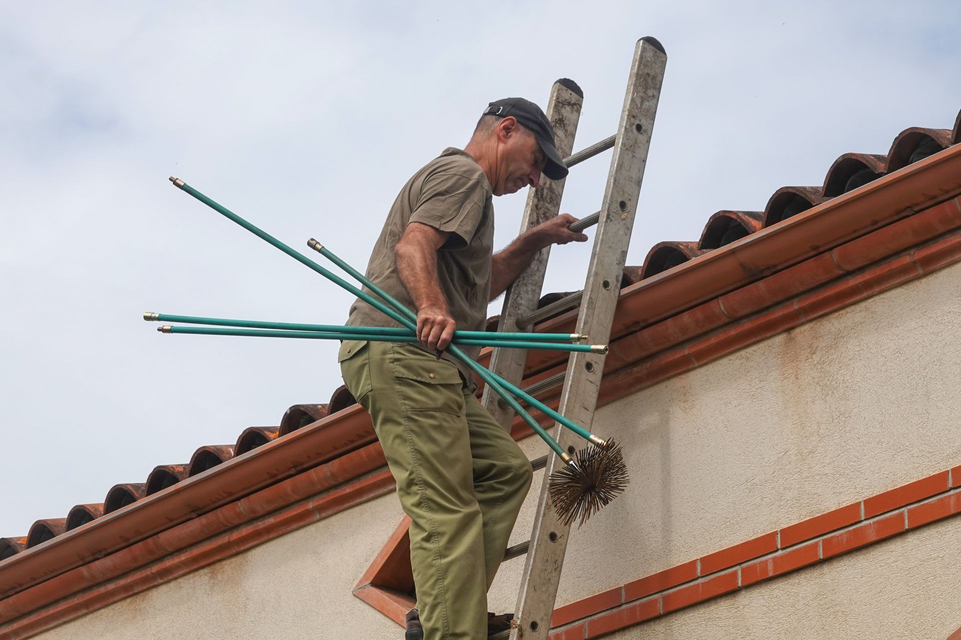 Man climbing ladder onto a roof, holding chimney cleaning rods and brush. Man climbing ladder onto a roof, holding chimney cleaning rods and brush.