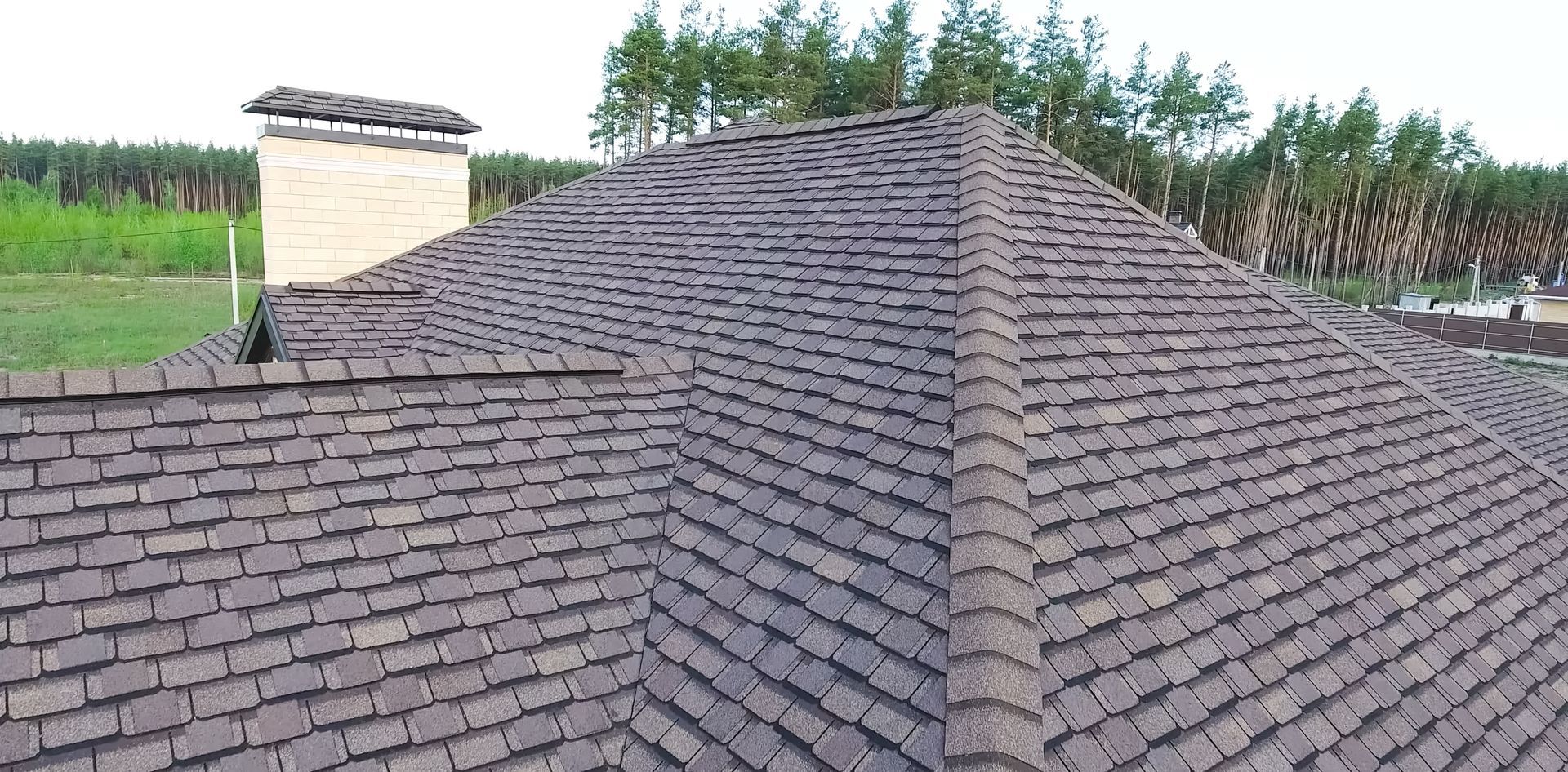 A brown shingled roof of a building with a chimney and trees in the background.