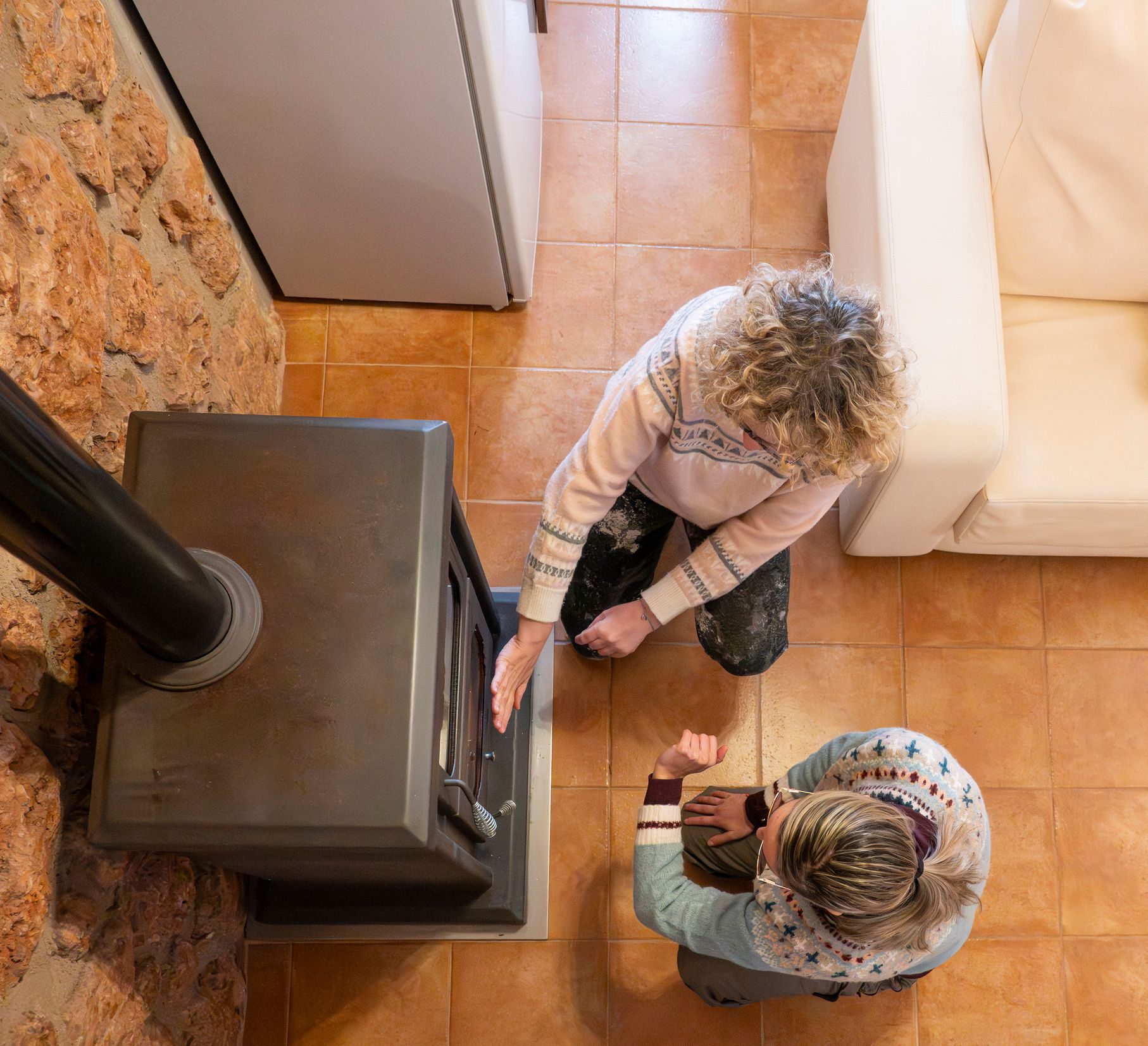 Two people kneeling by a dark stove, one touching it; tiled floor, stone wall, white couch. Two people kneeling by a dark stove, one touching it; tiled floor, stone wall, white couch.