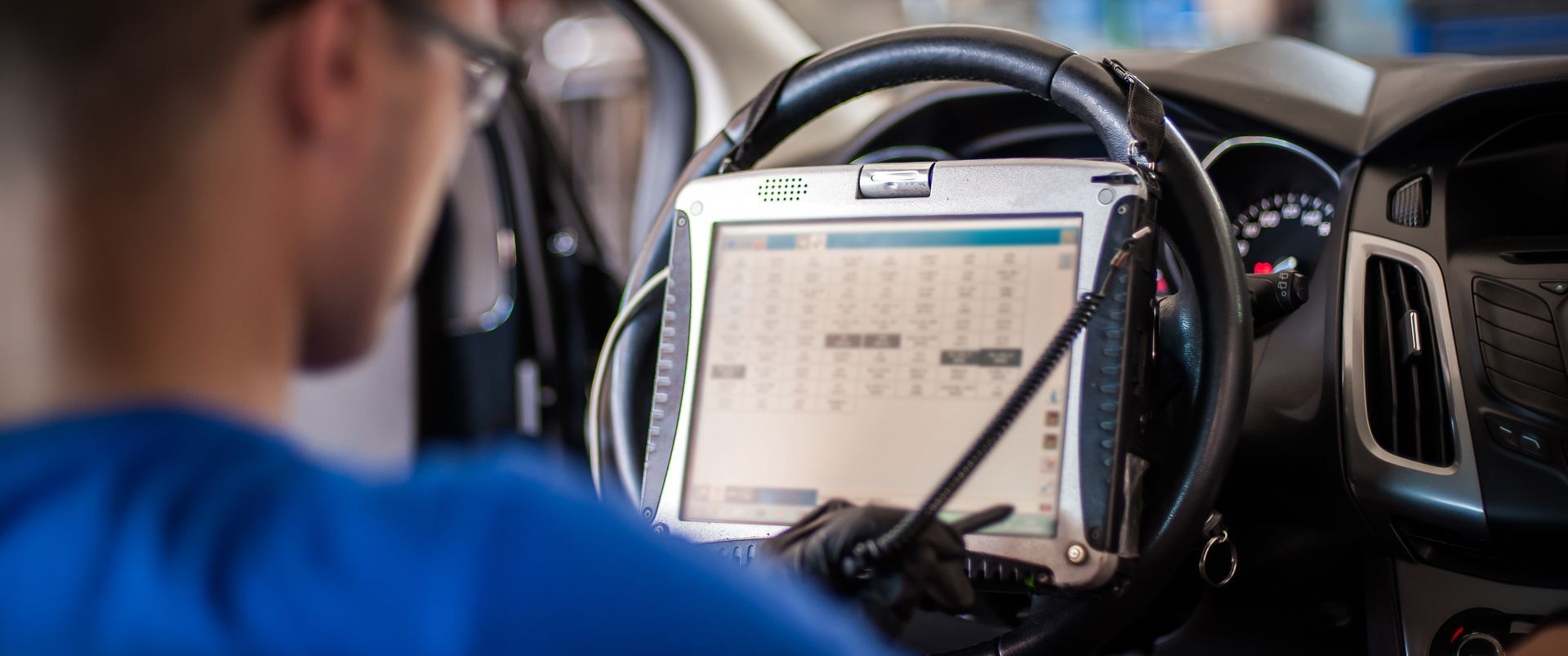Mechanic using a diagnostic tablet in a car. They wear glasses and a blue shirt, working in the vehicle's interior.
