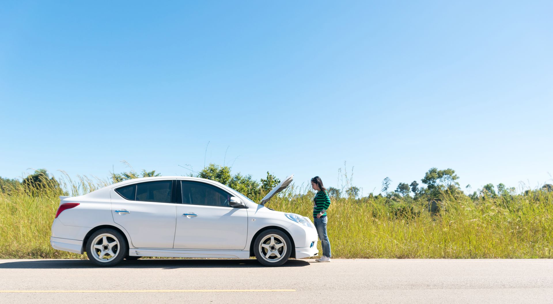 White sedan parked roadside with a person standing beside the front door in a sunny grassy area.