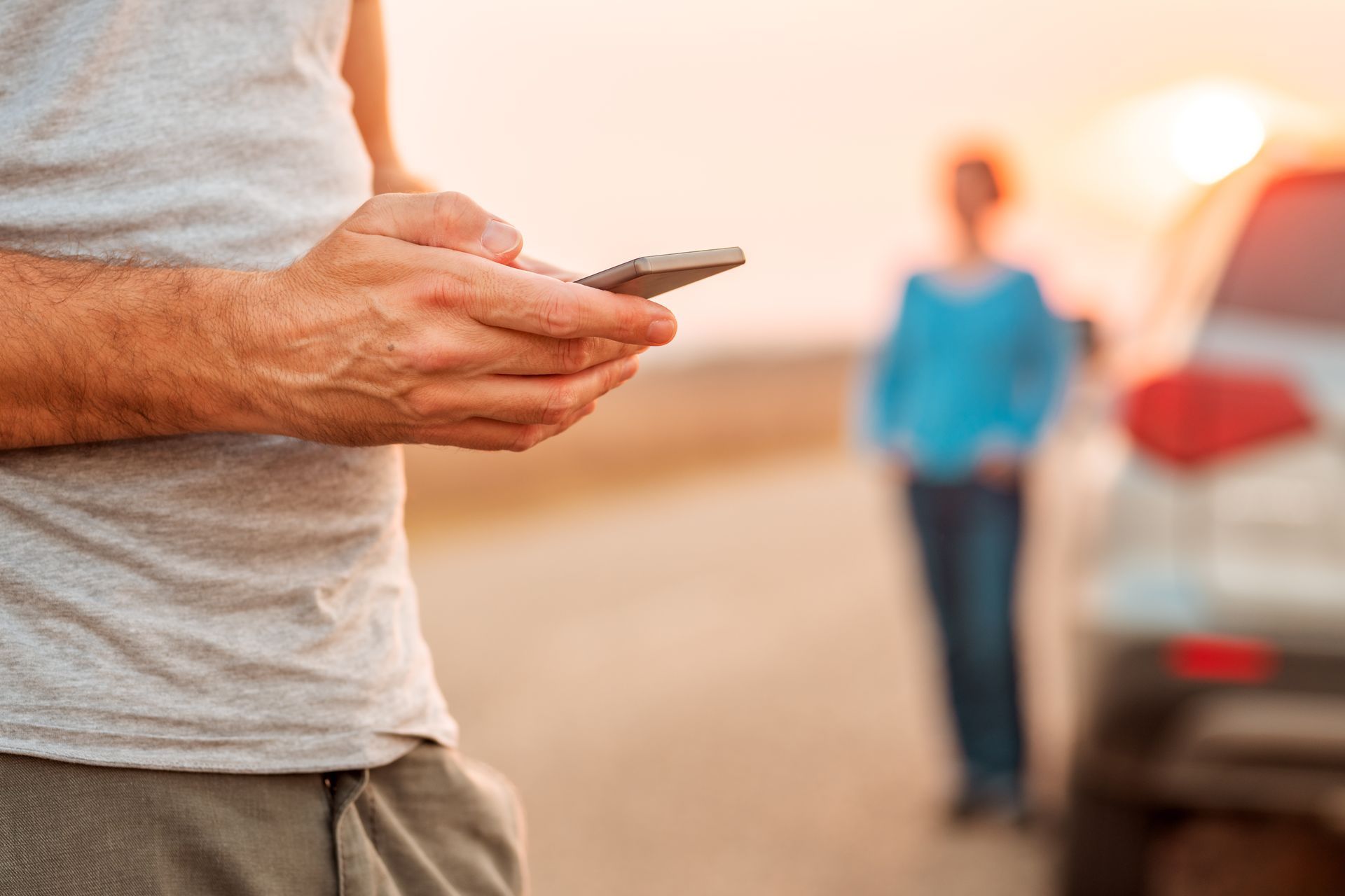 Person holding a smartphone beside a parked car on a roadside, with another person blurred in the background Person holding a smartphone beside a parked car on a roadside, with another person blurred in the background