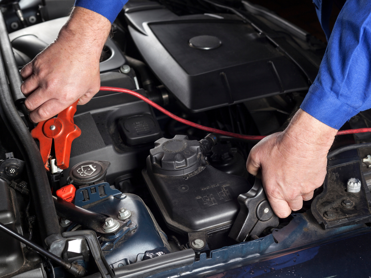 Mechanic's hands attaching jumper cables to a car battery in an engine bay; red and black cables.