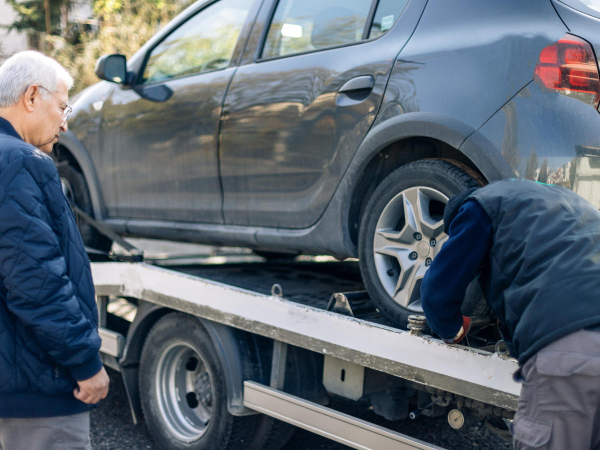 A gray car being loaded onto a tow truck. Man watches as worker secures the vehicle. Outdoors.