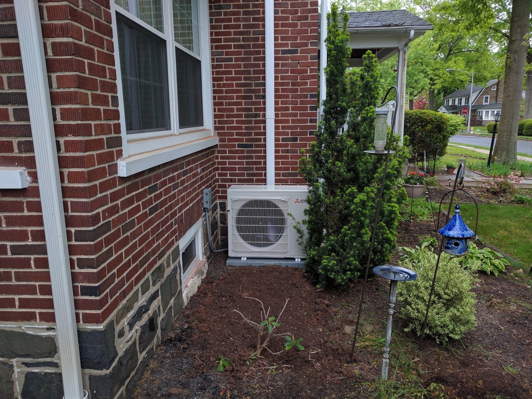 Exterior of a brick building with an air conditioning unit; a bush and birdhouse are nearby.