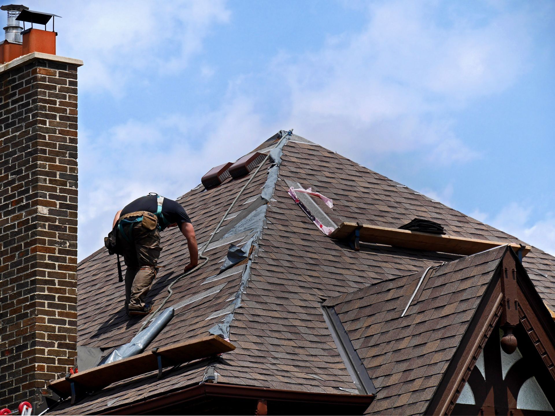 Roofing worker repairing shingles on a house roof next to a chimney under a blue sky.