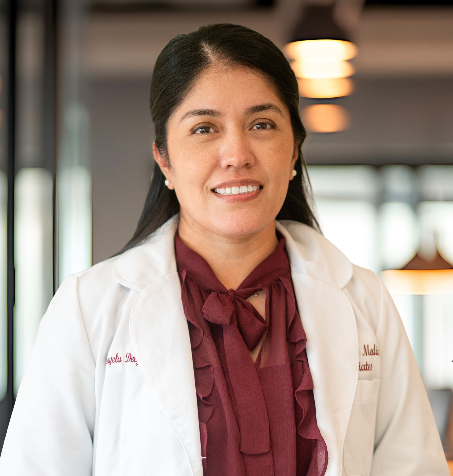 Woman in a white lab coat and burgundy blouse, smiling in a bright indoor setting.