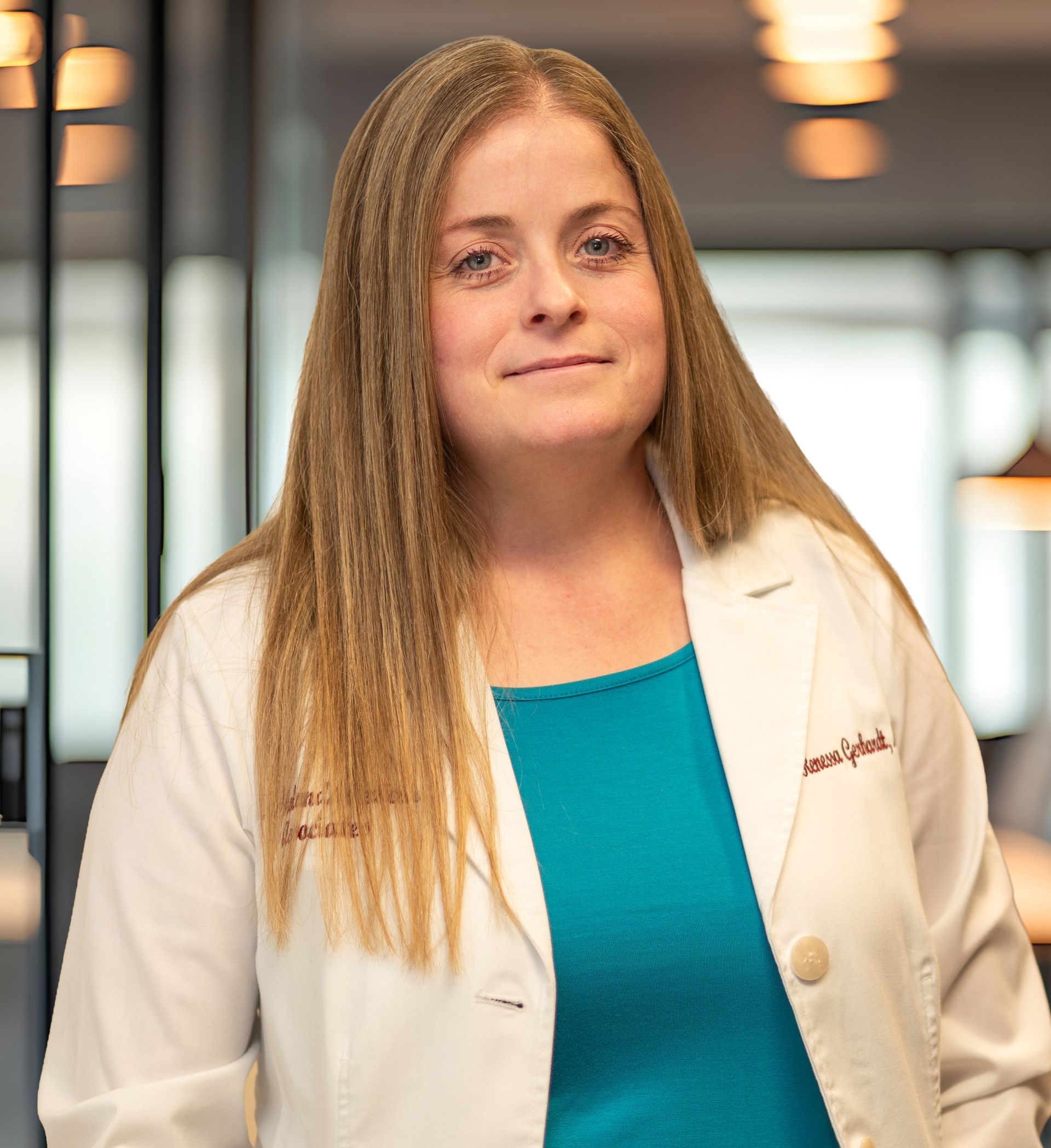 Woman in a white lab coat and teal shirt standing in a hallway with blurred lights behind her
