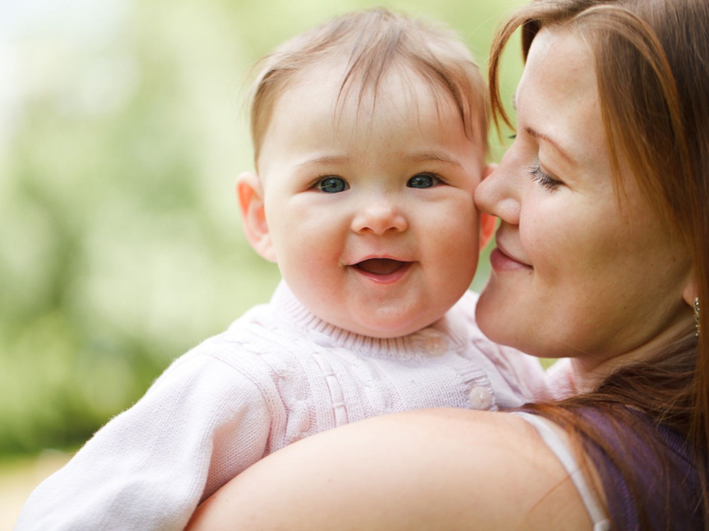 Smiling baby held close by an adult outdoors with a soft green background