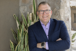 Smiling person with glasses wearing a navy blazer and lavender button-down shirt, standing with arms crossed by a plant.