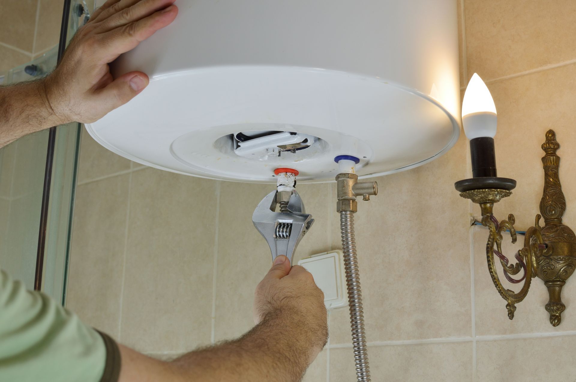 Person using a wrench on plumbing beneath a white water heater in a bathroom.