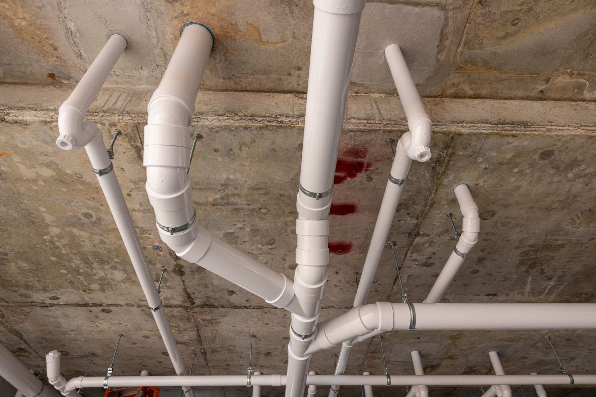 White PVC pipes and fittings running along a concrete ceiling, likely a plumbing system installation.