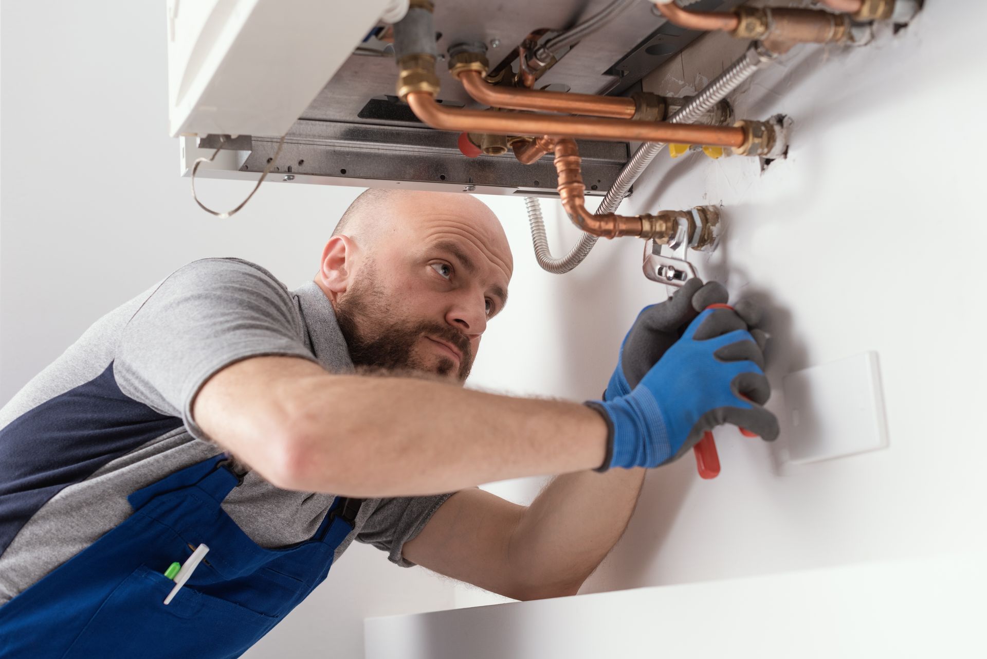Plumber repairs a boiler. He uses pliers, wearing blue gloves, in a white room.