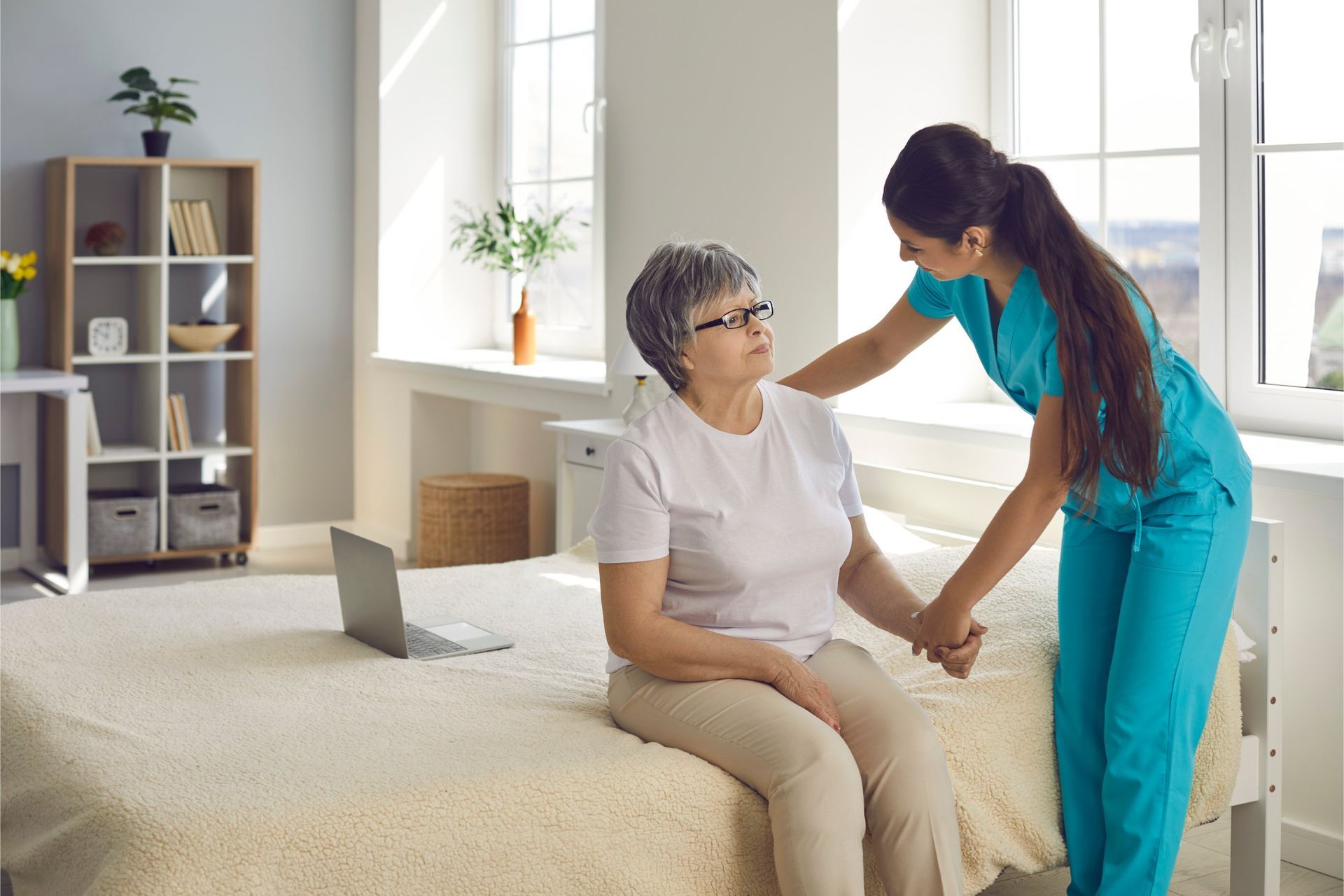 Nurse assisting an older adult woman to stand, in a bedroom with a laptop and window.