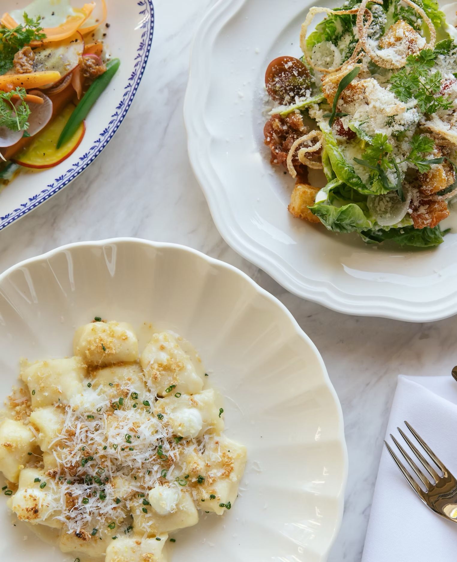Three plated gourmet dishes on a white table, including gnocchi and herb-topped pasta.