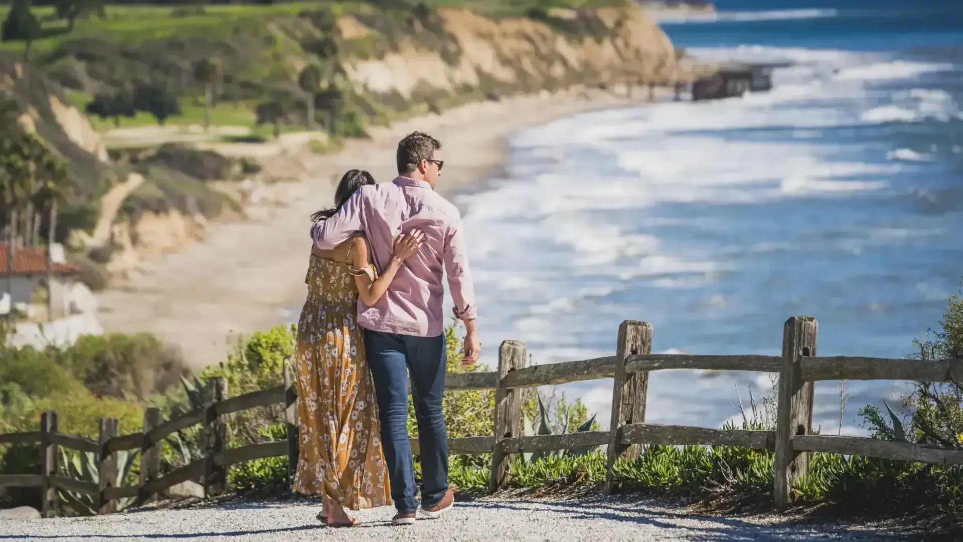 A couple stands with their arms around each other, looking at the ocean from a cliffside path behind a wooden fence.