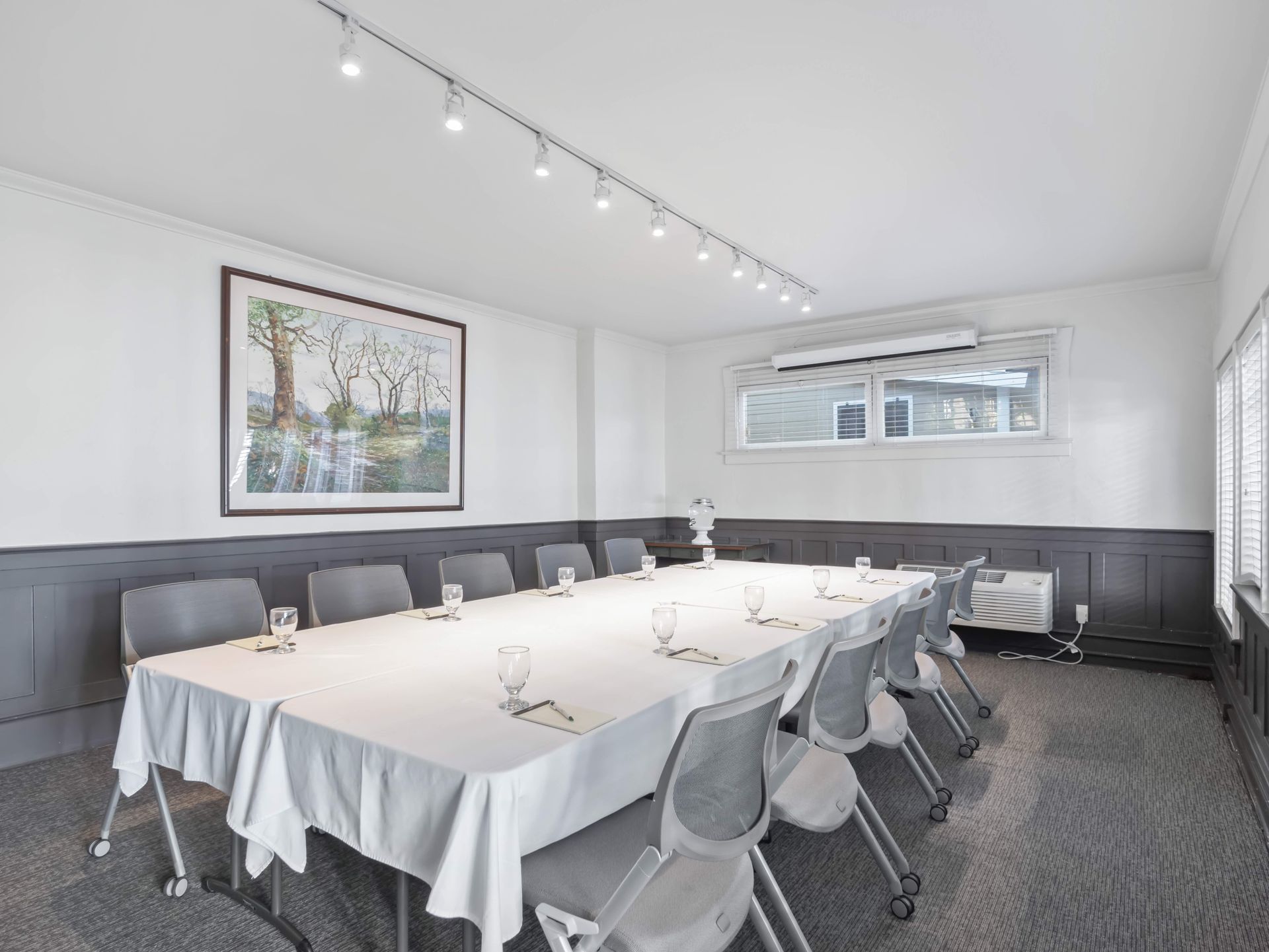 A conference room with a long table covered in a white cloth, set with glasses and grey chairs under track lighting.