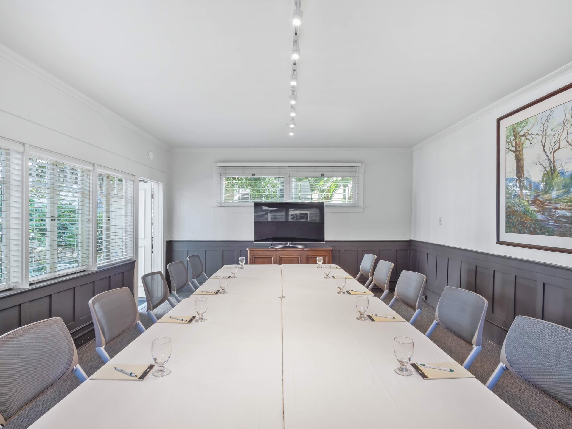 A conference room with a long white table, gray chairs, track lighting, windows with blinds, and a wall-mounted television.