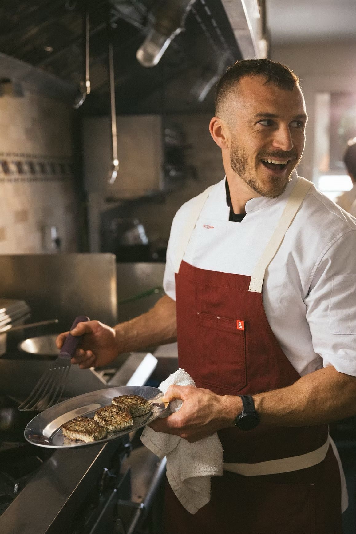 Chef in a red apron cooking in a restaurant kitchen, smiling while holding a skillet.