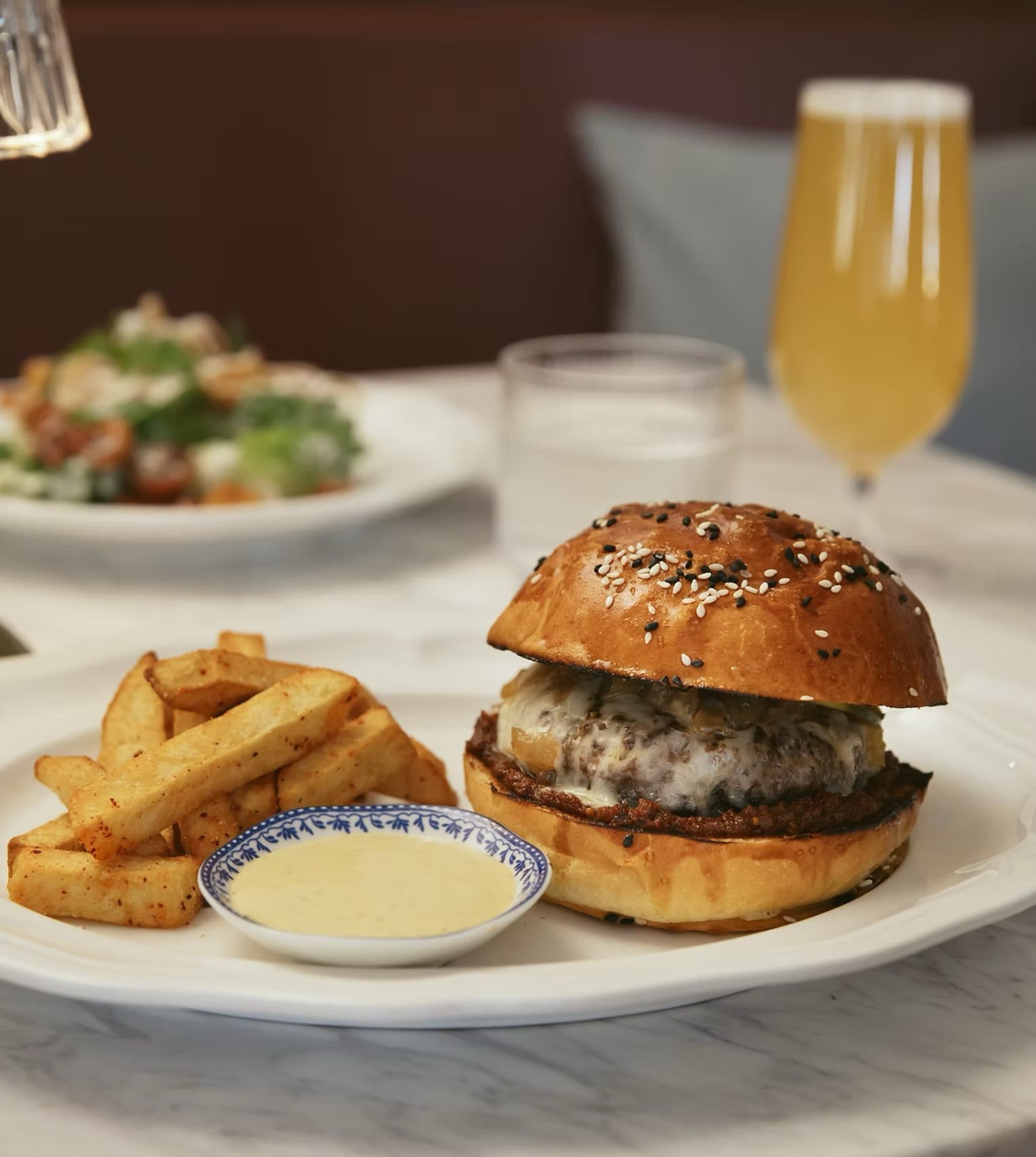 Burger with sesame bun, fries, dipping sauce, and a pale beer on a restaurant table.