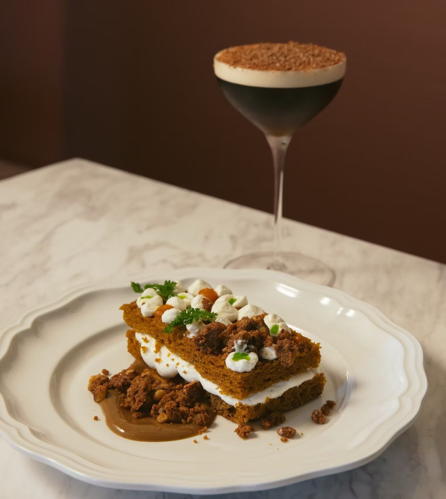 Dessert on a white plate beside a chocolate-topped cocktail on a marble table