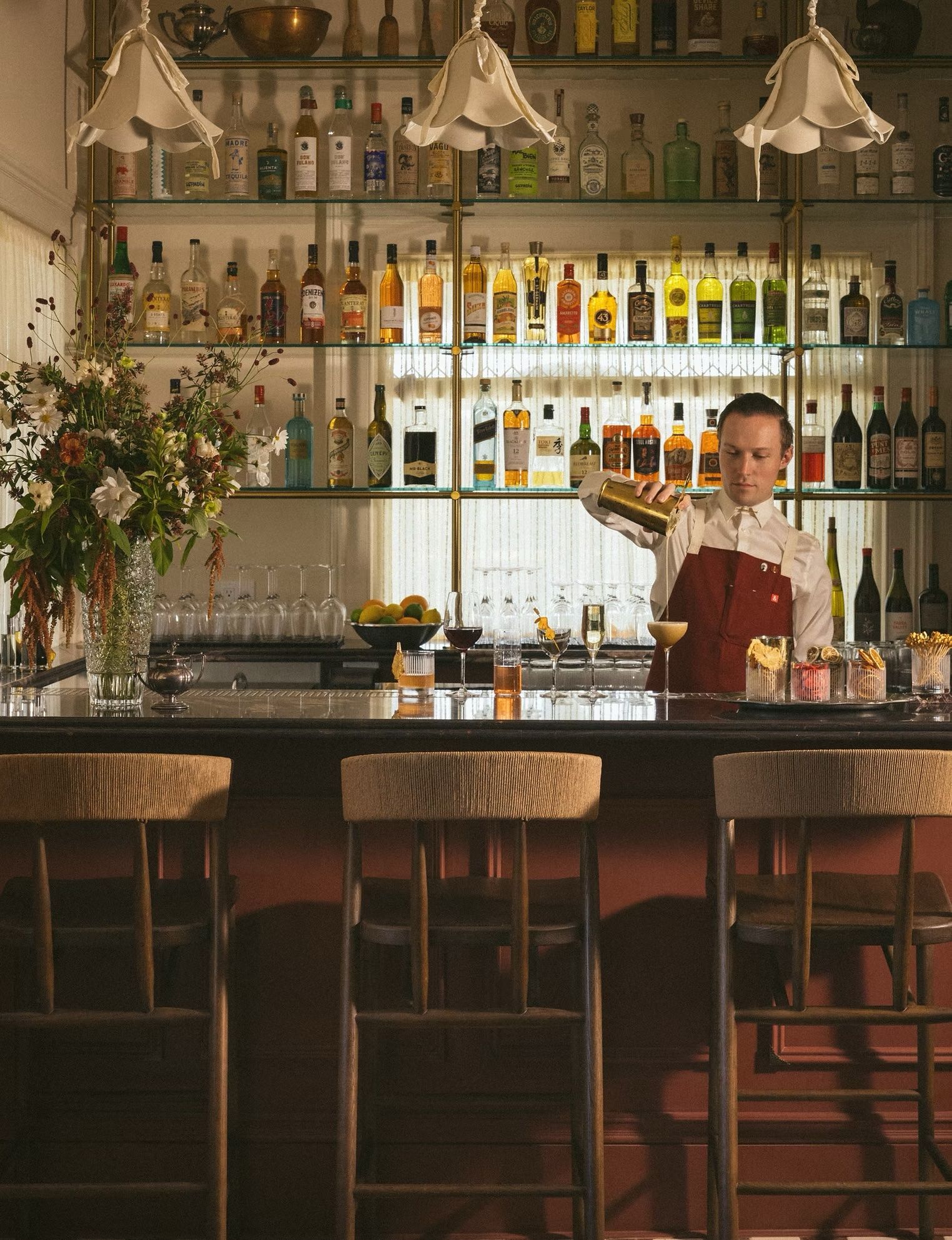 Bartender behind a bar in a warmly lit liquor-filled lounge, holding a tray of drinks.
