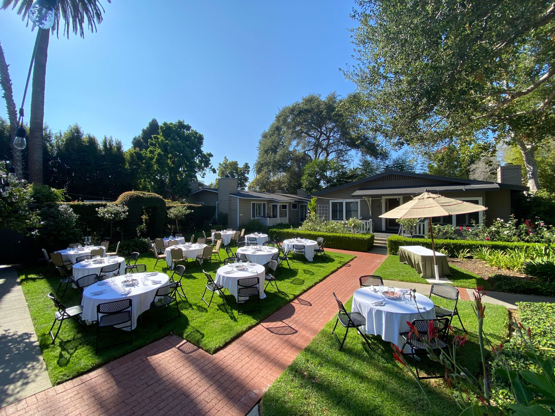 Outdoor garden space with white-clothed round tables set for a meal, arranged along a brick path near a house.