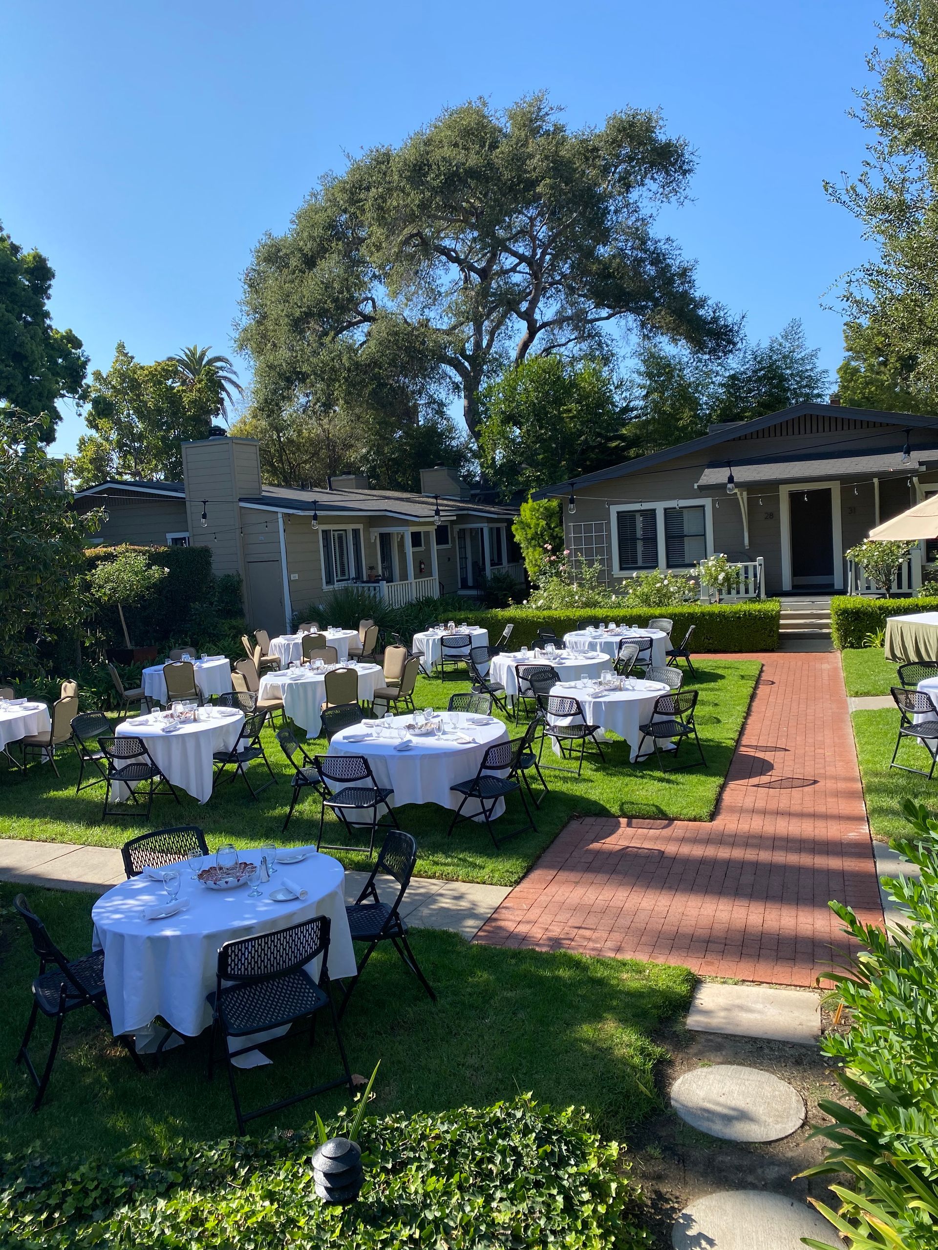 Outdoor restaurant patio with tables covered in white tablecloths on a grassy lawn, leading to small buildings.