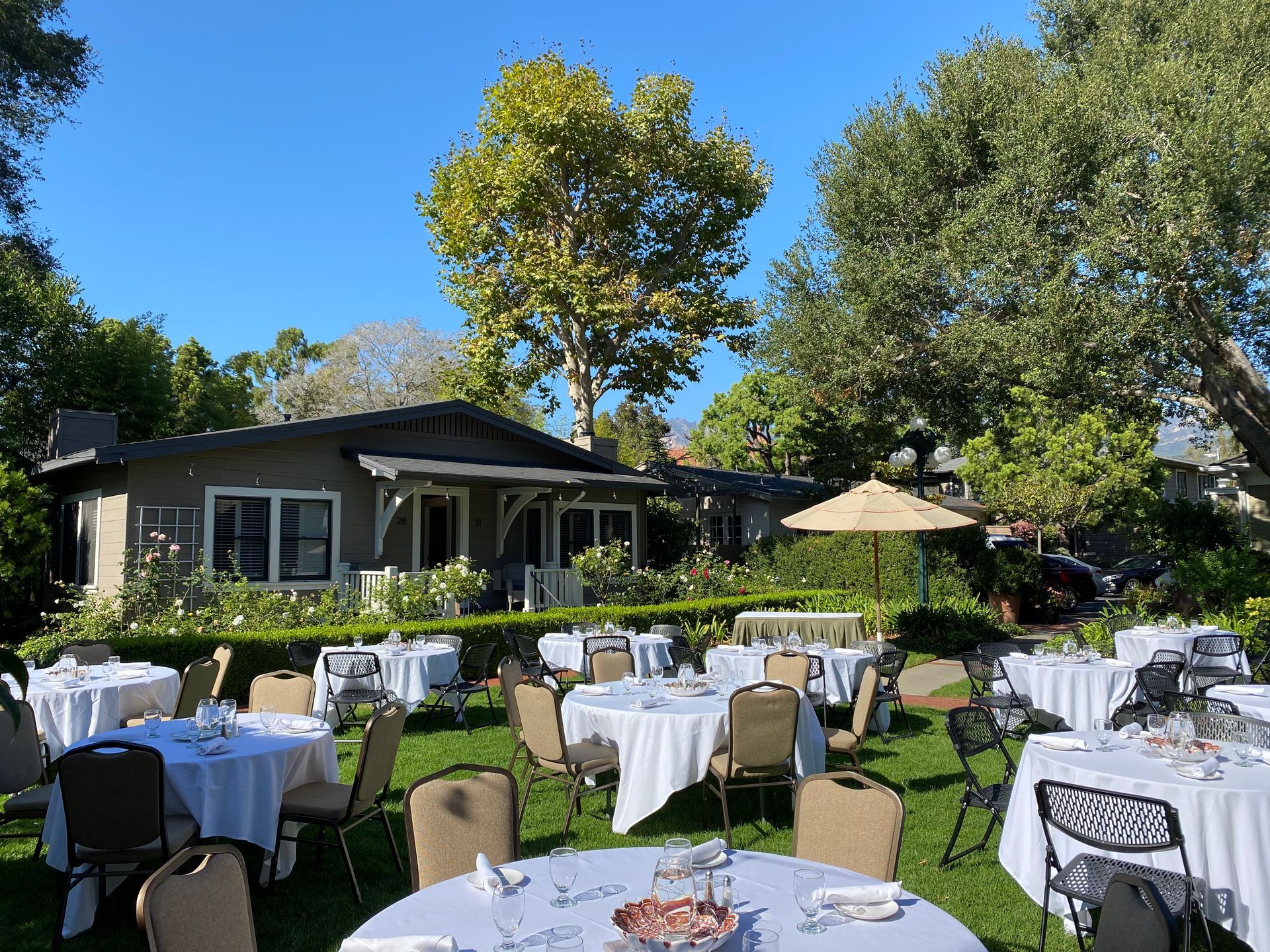 Outdoor seating area with round tables covered in white tablecloths on a grassy lawn in front of a house.