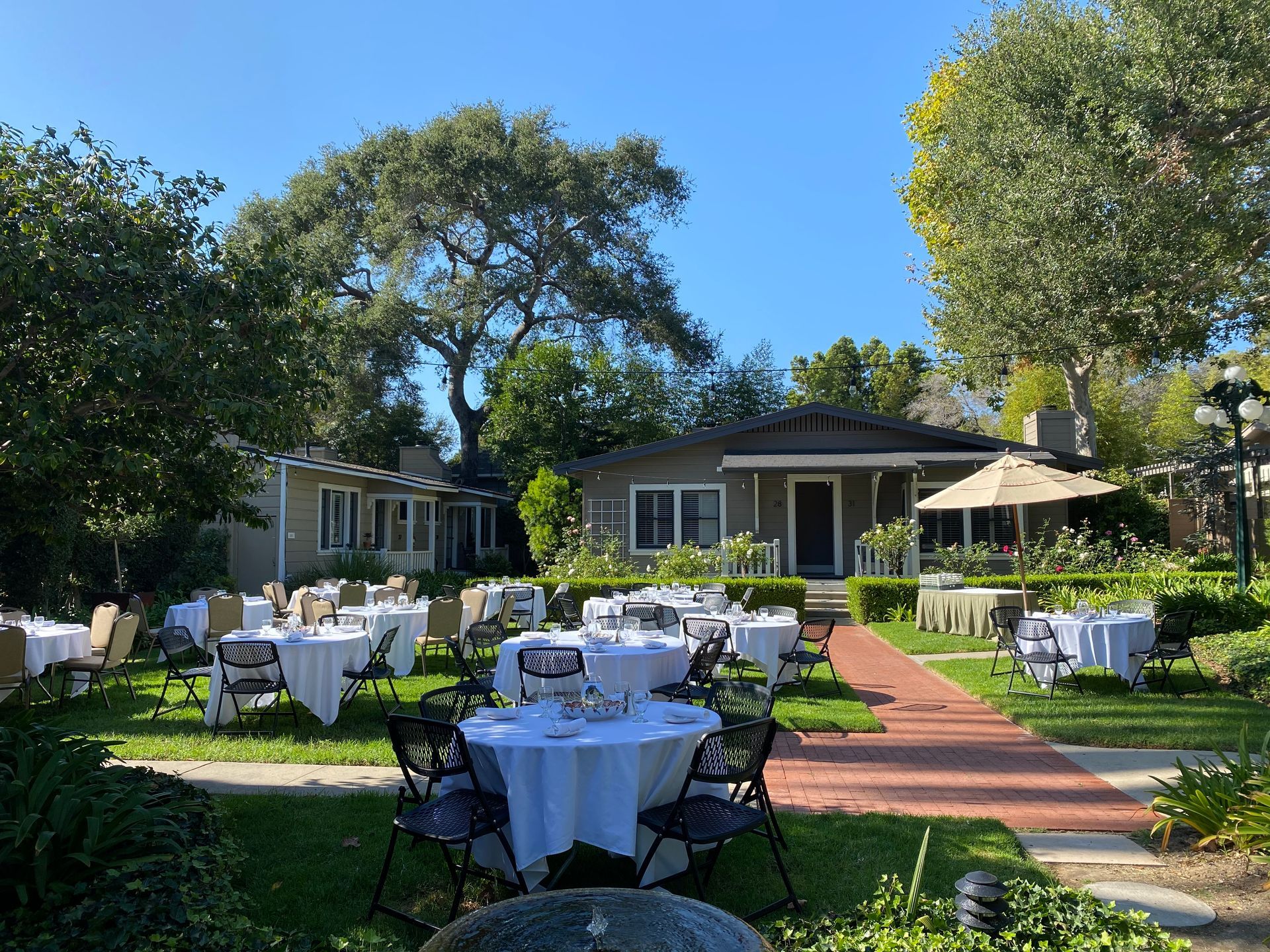 Outdoor dining setup on a grassy lawn with round tables covered in white tablecloths, chairs, and a brick path.
